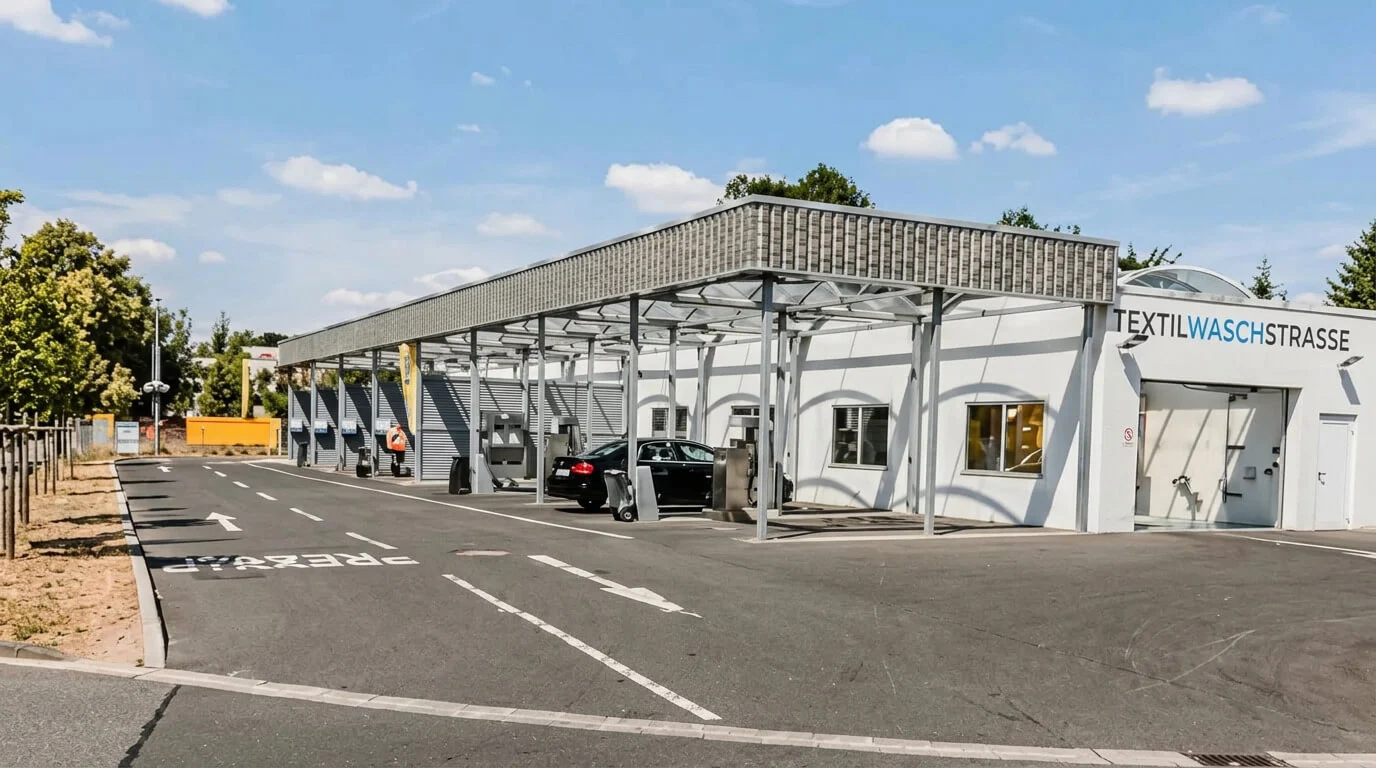 Eine Durchfahrtswaschanlage mit Autos und Menschen unter einem Metallvordach, darüber der blaue Himmel. Eine Durchfahrtswaschanlage mit Autos und Menschen unter einem Metallvordach, darüber der blaue Himmel.